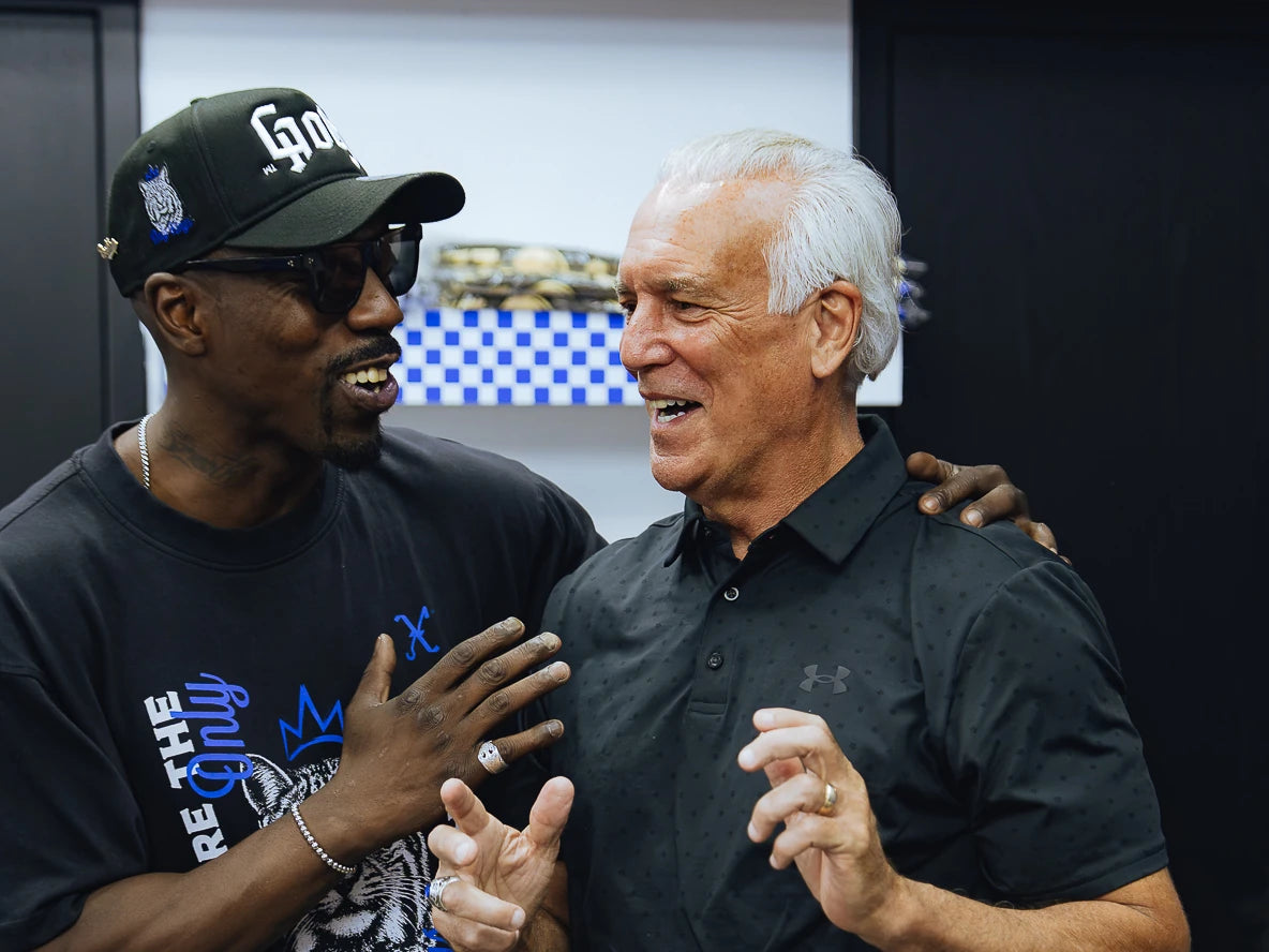 Two adults smiling and gesturing together at a barbershop, one in a GOTH cap and tee.